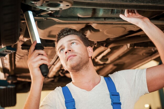 Jetzt sehe ich das Problem. Niedriger Blickwinkel eines selbstbewussten jungen Mannes in einheitlicher Haltelampe, während er in der Werkstatt unter einem Auto steht