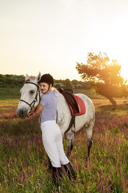 Jockey joven acariciando y abrazando al caballo blanco en el atardecer. llamarada del sol, destello solar