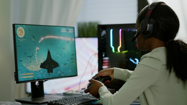 Foto jogador africano profissional jogando jogo de tiro espacial online em um computador poderoso usando controlador sem fio e fone de ouvido. mulher competidora cibernética realizando torneio de videogame usando joystick profissional