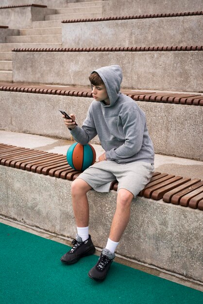 Jogador de basquete adolescente sentado sozinho em um campo de esportes usando um capuz usando um smartphone