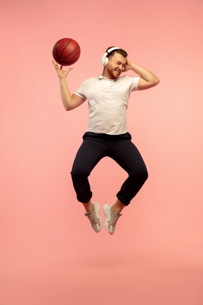 Jogador de basquete em ação. retrato de corpo inteiro de jovem salto em altura isolado no fundo rosa do estúdio. modelo masculino caucasiano. copyspace. emoções humanas, expressão facial, conceito de esporte.