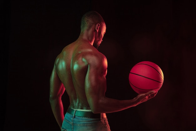 Jogador de basquete jovem musculoso afro-americano em ação de treinamento de jogabilidade, praticando em luzes de néon sobre o fundo escuro do estúdio. Conceito de esporte, movimento, energia, estilo de vida dinâmico e saudável.