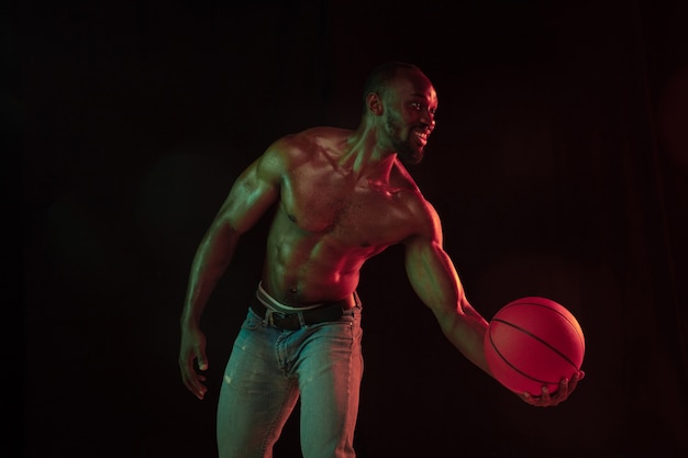 Jogador de basquete jovem musculoso afro-americano em ação de treinamento de jogabilidade, praticando em luzes de néon sobre o fundo escuro do estúdio. Conceito de esporte, movimento, energia, estilo de vida dinâmico e saudável.