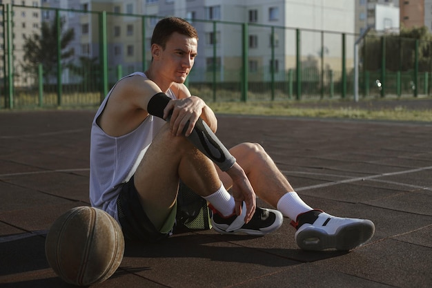 Foto jogador de basquete masculino sentado com bola na quadra de verão e fazendo uma pausa durante o treinamento