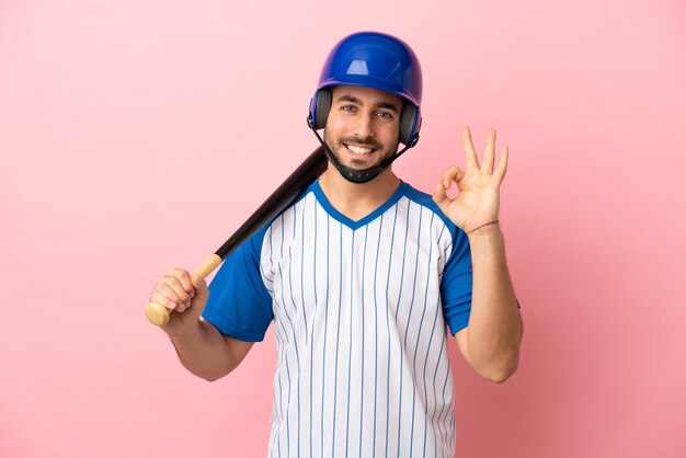 Jogador de beisebol com capacete e taco isolado em fundo rosa mostrando sinal de ok com os dedos