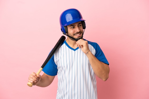 Jogador de beisebol com capacete e taco isolado em um fundo rosa olhando para o lado e sorrindo