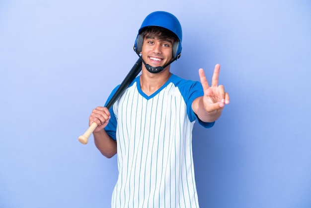 Jogador de beisebol homem caucasiano com capacete e taco isolado em fundo azul sorrindo e mostrando sinal de vitória