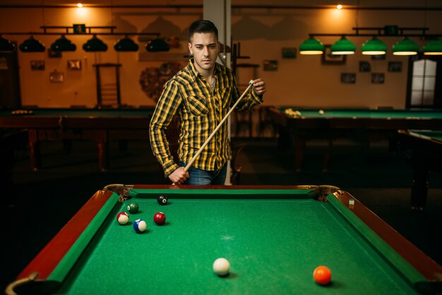 Jogador de bilhar masculino com taco à mesa com bolas coloridas. Homem joga sinuca americana no bar esportivo