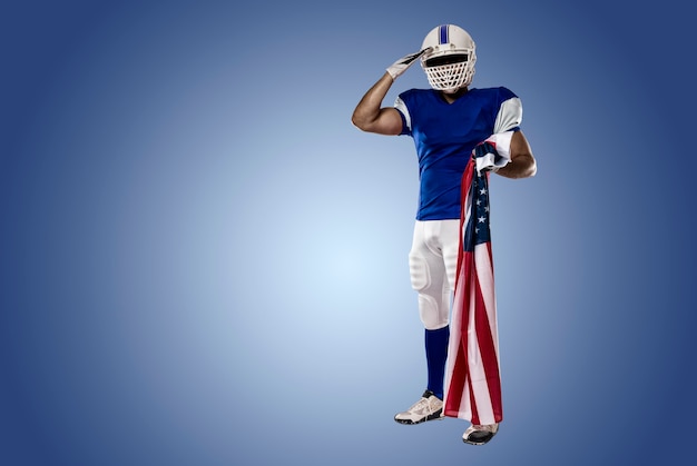 Jogador de futebol com uniforme azul saudando com bandeira americana, em parede azul