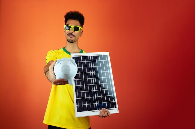 Jogador de futebol homem negro celebrando segurando painel solar fotovoltaico e moeda de porco isolada em fundo laranja