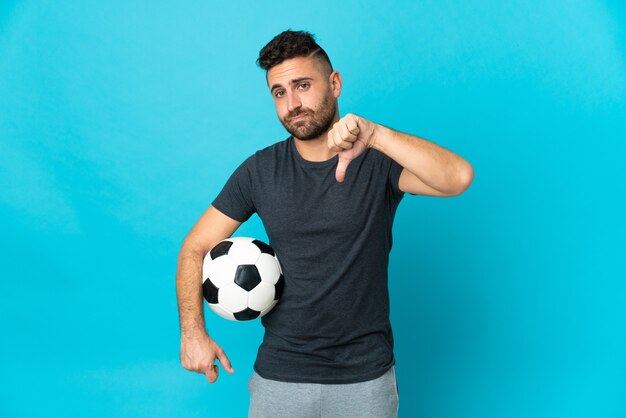 Jogador de futebol isolado em fundo azul mostrando o polegar para baixo com expressão negativa