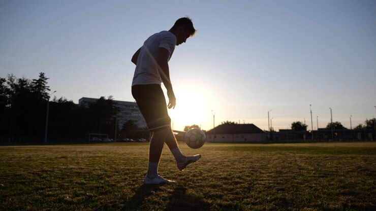 Foto jogador de futebol profissional fazendo malabarismo com uma bola de futeblol no estádio ao pôr do sol jovem a chutar a bola no campo verde desportista a praticar truques no prado com a luz do sol no fundo conceito de futebil freestyle