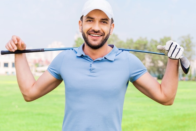 Jogador de golfe profissional confiável. Jovem sorridente carregando o taco de golfe nos ombros em pé no campo de golfe