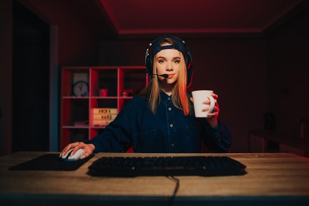 Foto jogador de menina em um fone de ouvido e com um copo de bebida na mão joga jogos online no computador à noite