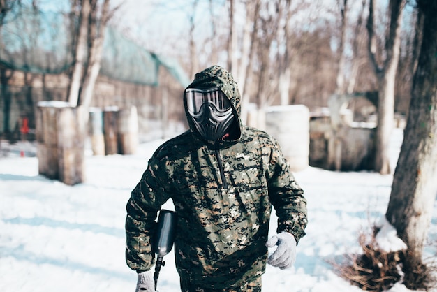 Jogador de paintball masculino com máscara de proteção e uniforme detém a arma do marcador nas mãos, soldado antes da batalha na floresta de inverno. Esporte radical, equipamento de jogo militar