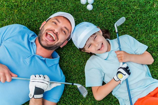 Jogadores de golfe felizes. Vista superior de um menino alegre e seu pai segurando tacos de golfe e sorrindo enquanto estava deitado na grama verde