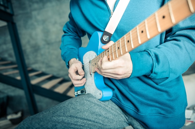 Jogando ativamente. Músico profissional vestindo um moletom brilhante carregando uma guitarra eletrônica azul enquanto está sozinho em casa