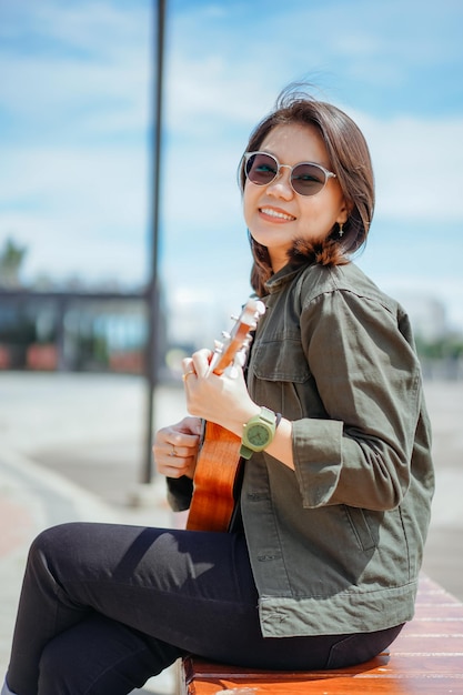 Jogando o Ukulele de uma jovem bela mulher asiática vestindo jaqueta e calça jeans preta posando ao ar livre
