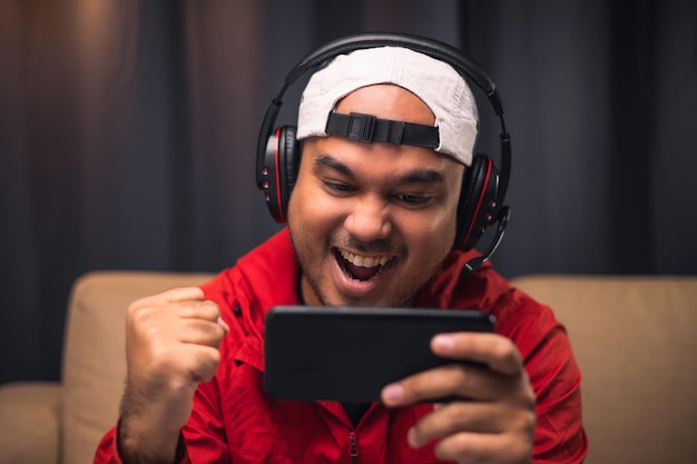 Jogando videogame no smartphone Jovem asiático bonito sentado no sofá segurando o celular na sala de estar Felicidade Streamer Homem indiano usando fone de ouvido jogando jogo online na câmara escura