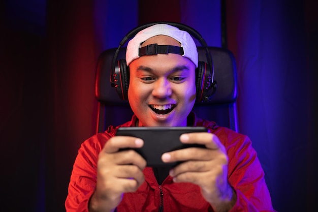 Foto jogando videogame no smartphone. jovem bonito asiático sentado na cadeira segurando o celular na sala de estar. felicidade streamer homem indiano usando fone de ouvido jogando jogo online na câmara escura.