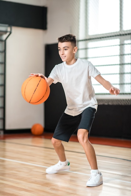 Jogar basquete. Adolescente de camiseta branca jogando basquete na academia e parecendo envolvido