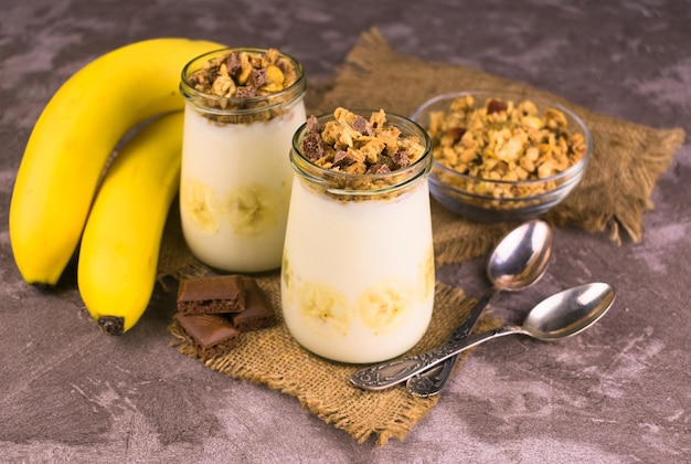 Joghurt mit Banane und Müsli in Gläsern auf grauem Hintergrund.