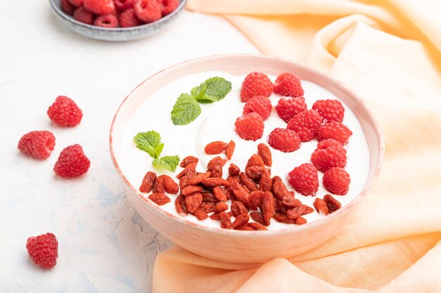 Joghurt mit Himbeere und Goji-Beeren in Keramikschale auf weißem Betonhintergrund Seitenansicht