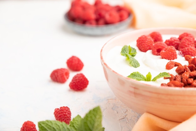 Joghurt mit Himbeeren und Goji-Beeren in Keramikschale auf weißem Betonhintergrund und orangefarbenem Leinengewebe. Seitenansicht, Nahaufnahme, selektiver Fokus.