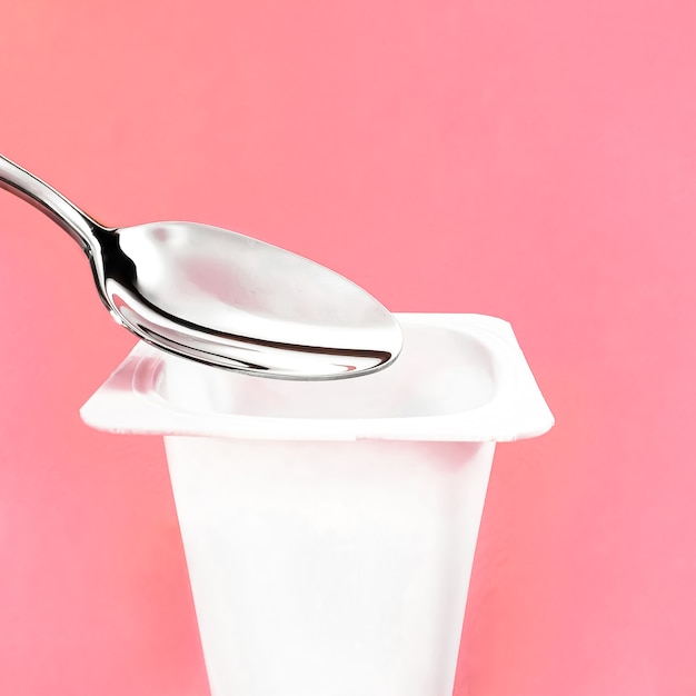 Joghurtbecher und silberner Löffel auf rosafarbenem Hintergrund, weißer Kunststoffbehälter mit Joghurtcreme, frischem Milchprodukt für gesunde Ernährung und ausgewogene Ernährung