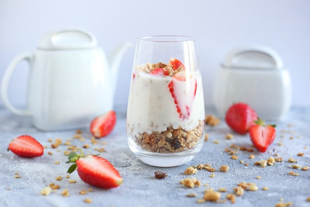 Joghurtdessert mit Haferflocken, Walnuss und Erdbeeren auf hellgrauem Tisch mit Teeservice und halben Erdbeeren. Gesundes Essen.