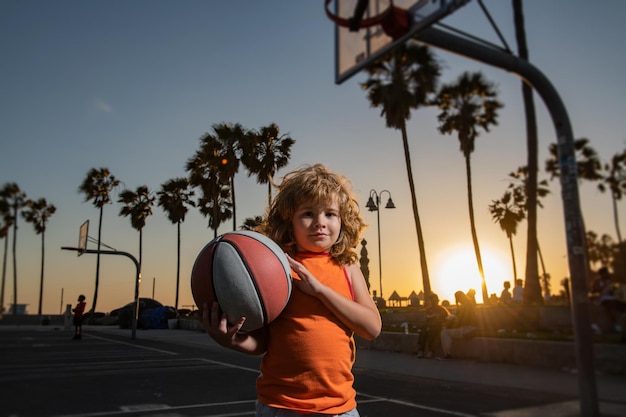 Jogo de basquete infantil. Menino bonitinho segurando uma bola de basquete, tentando fazer uma pontuação. Adorável criança jogando basquete na quadra no pôr do sol.