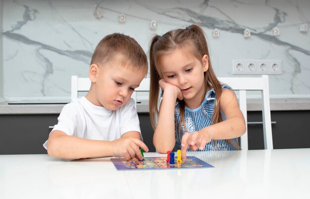 Jogo de tabuleiro e conceito de lazer infantil. as crianças se divertem, riem e se divertem jogando jogo de tabuleiro. elas seguram figuras nas mãos, fichas vermelhas, azuis, verdes. menino e menina jogando jogo de tabuleiro em casa