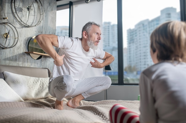 Jogo, observação. Pai adulto barbudo com roupas de casa fazendo movimento e fazendo careta enquanto está sentado na cama e a criança assistindo