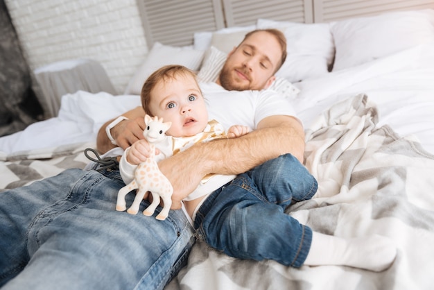 Jogos ativos. Pai atencioso e bonito, atlético, deitado na cama em casa, segurando a criança nos braços enquanto seu filho brinca com o brinquedo
