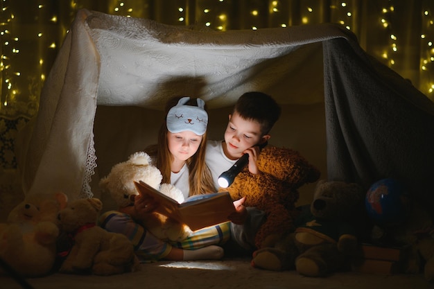 Jogos de leitura e família na tenda infantil menino e menina com livro e lanterna antes de ir para a cama