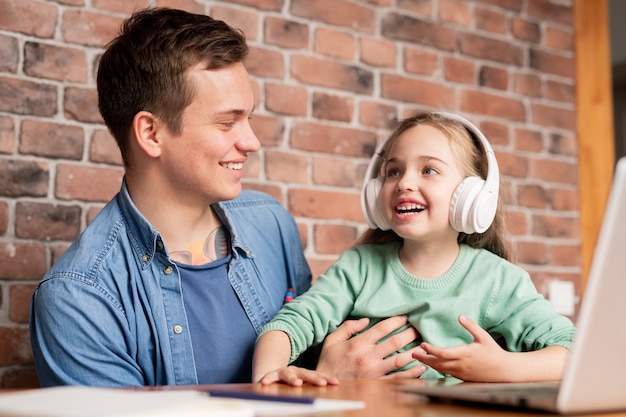 Jolly emocionada hija en auriculares inalámbricos sentado a la mesa con el padre y escuchando la música preferida por papá