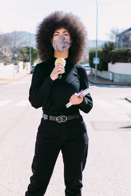 Jornalista repórter de televisão com cabelo afro, usando máscara de pele de leopardo para se proteger de um vírus, relatando matéria na rua