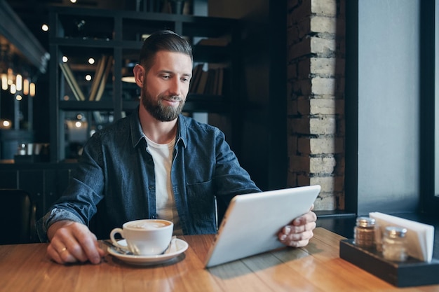 Jovem a beber café num café e a usar um tablet. Trabalhador independente. Lugar de trabalho. Almoço