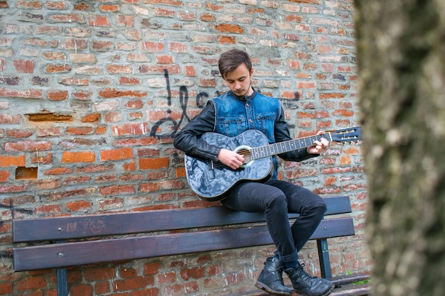 Jovem a tocar guitarra sentado num banco contra uma parede de tijolos