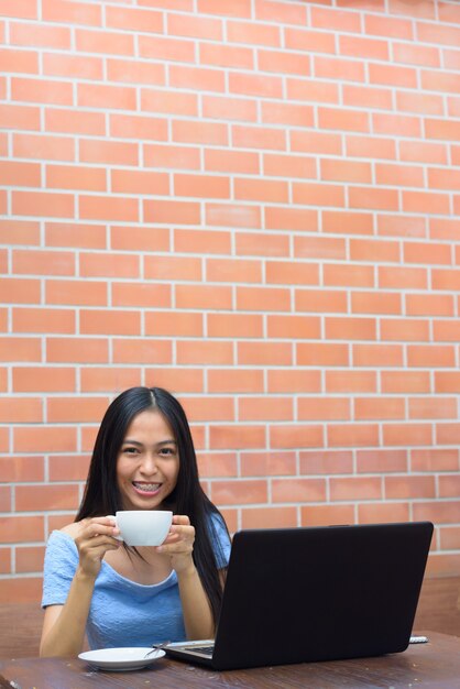 Jovem adolescente asiática feliz bebendo cappuccino com o laptop na mesa de madeira