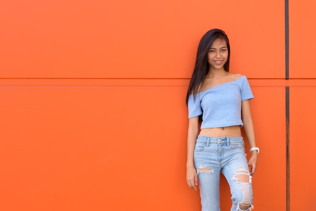 Jovem adolescente asiática feliz sorrindo enquanto se inclina contra uma parede pintada de laranja