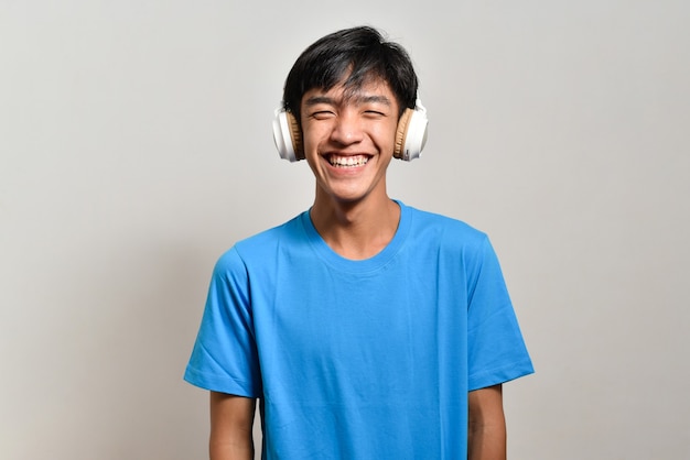 Jovem adolescente asiático feliz ouvindo música com seus fones de ouvido sobre uma parede cinza