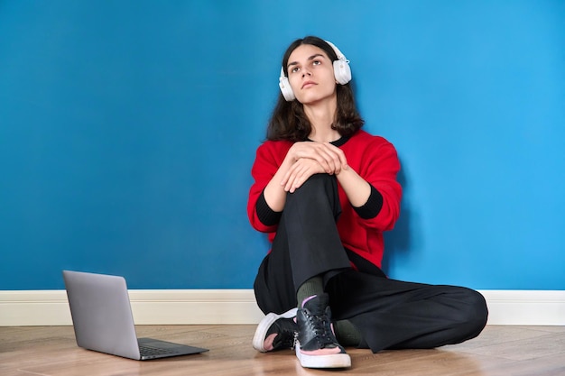 Jovem adolescente em fones de ouvido com laptop sentado no chão em fundo azul
