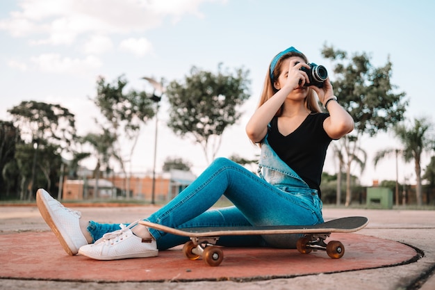 Foto jovem adolescente sentada em um skate ao ar livre, tirando fotos com uma câmera.