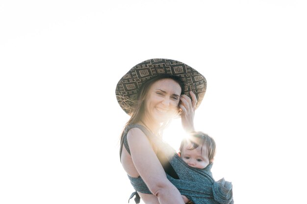 Jovem adulta com chapéu carregando seu bebê na praia no verão.