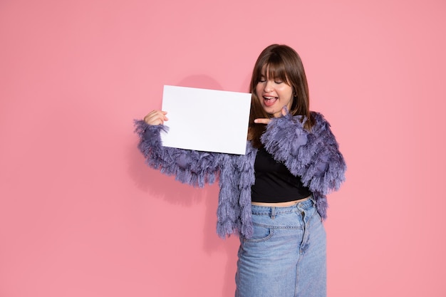 Jovem adulta de cabelos castanhos em pé e mostrando com o dedo no fundo rosa com quadro branco em branco para assinar no fundo rosa no estúdio. surpreenda, animado. retrato