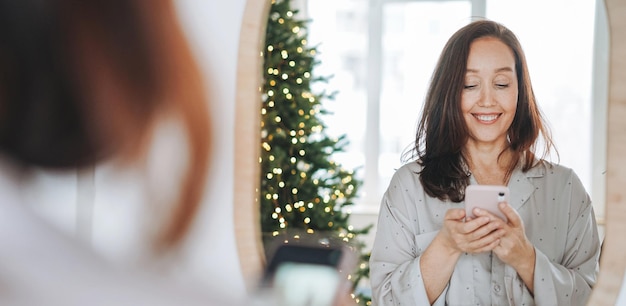 Jovem adulta de quarenta anos de meia idade linda mulher com cabelos escuros de pijama aconchegante olhando no espelho e tomando selfie na manhã de natal no quarto em casa