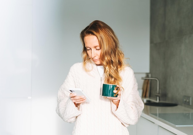 Jovem adulta de quarenta anos linda mulher loira com cabelo comprido usando smartphone e bebendo café da manhã na cozinha em casa
