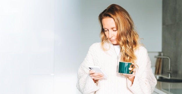 Foto jovem adulta quarenta anos linda loira com cabelos longos usando smartphone e tomando café da manhã na cozinha em casa