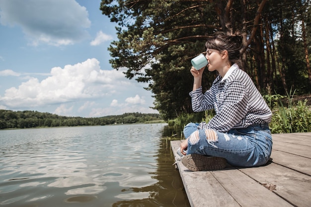 Jovem adulta sentada na doca de madeira tomando café e olhando para o lago